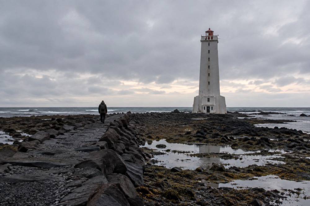 The Grotta lighthouse stands on a rocky shore under a grey sky with the sea stretching behind it.