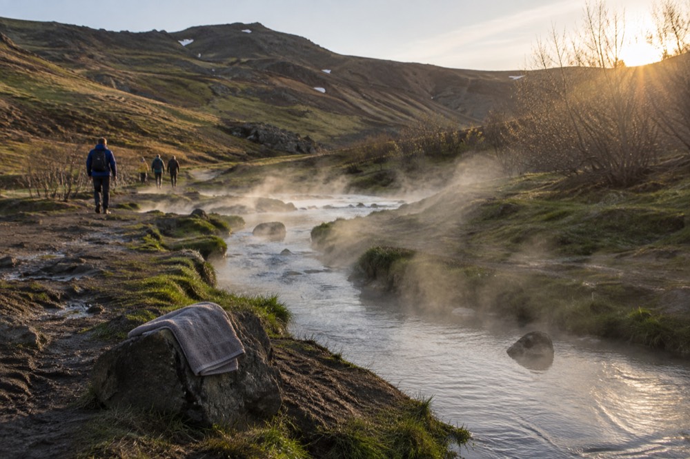 Steam rises from a hot river winding through a green valley with hikers on the banks.
