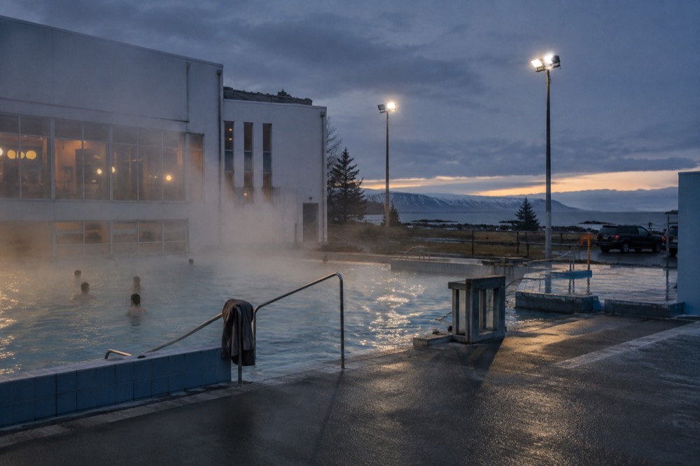 The exterior of a Reykjavik public swimming pool with geothermal steam rising from the water, surrounded by a simple low-rise building and a view of the sea in the distance, no people.