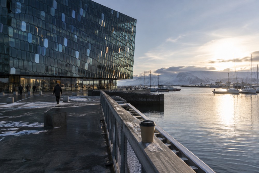 A modern concert hall with a faceted glass facade reflecting light, beside a harbour