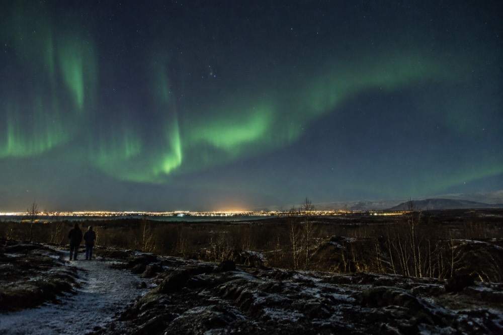 Green aurora bands over a dark hillside above Reykjavik city lights