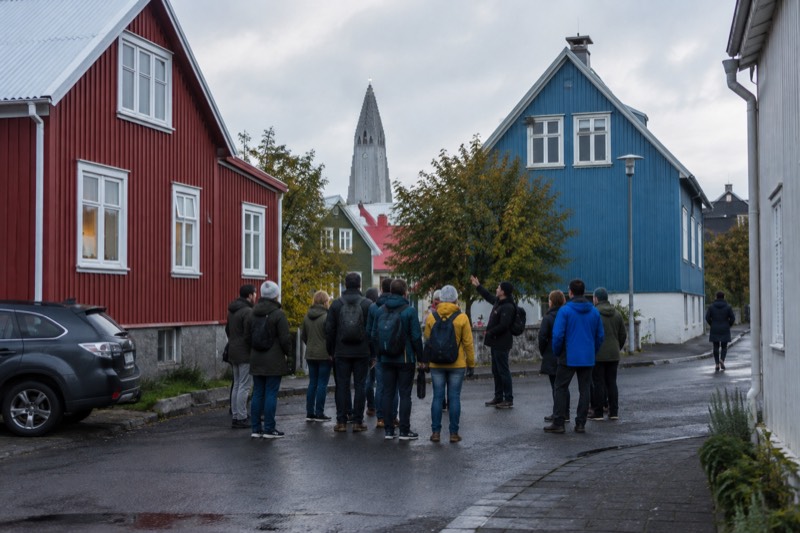 A free walking tour group paused on a Reykjavik street corner, the guide gesturing toward a stop on the route, low colourful houses and a soft northern sky filling the frame behind them.