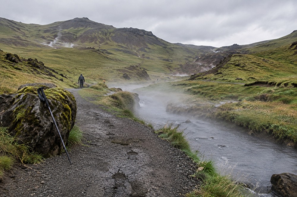 Steam rising from a geothermal stream beside a gravel hiking trail in a green valley