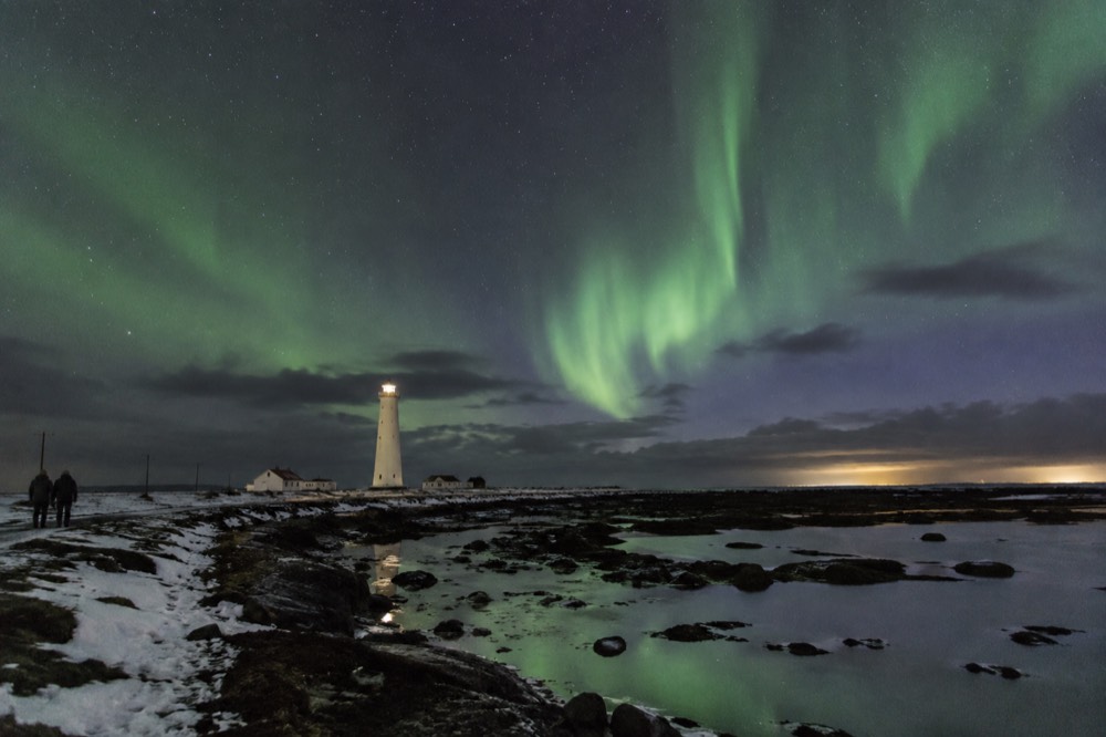 Green aurora over a dark coastline with a small lighthouse silhouette