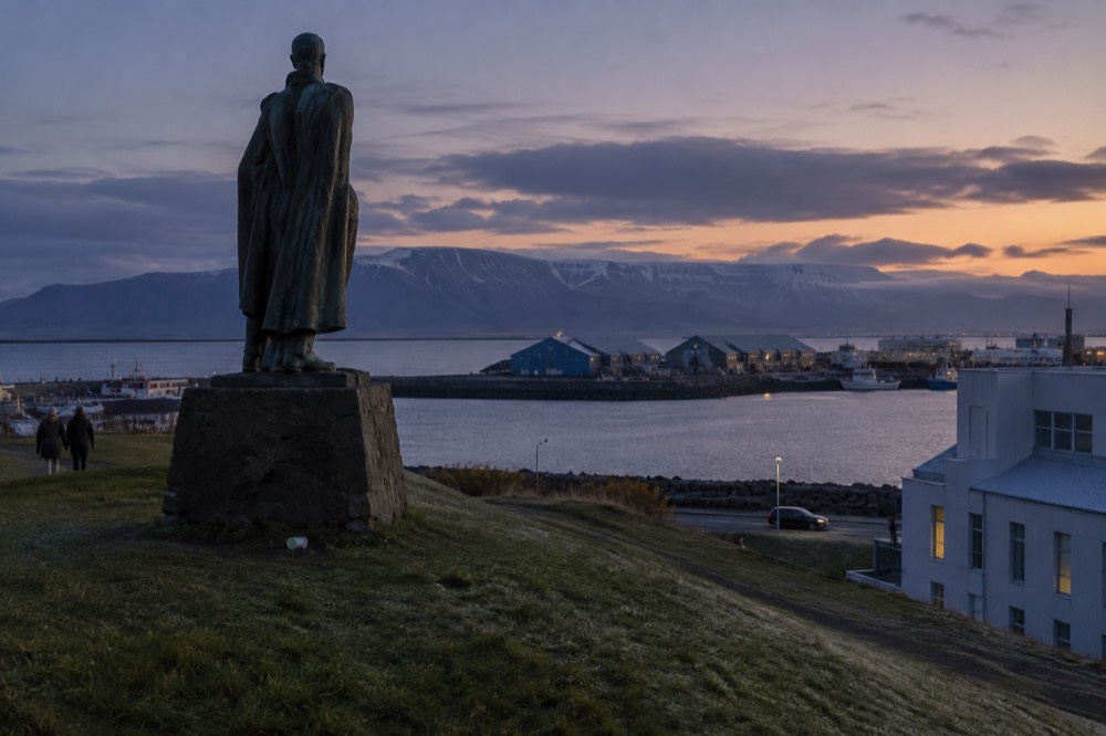 Statue on a grassy hill overlooking a harbour and distant mountains
