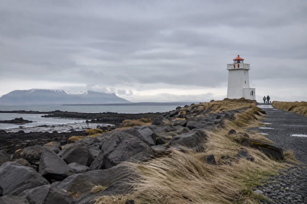 A squat white lighthouse standing on a low grassy island, with a gravel causeway stretching toward the shore