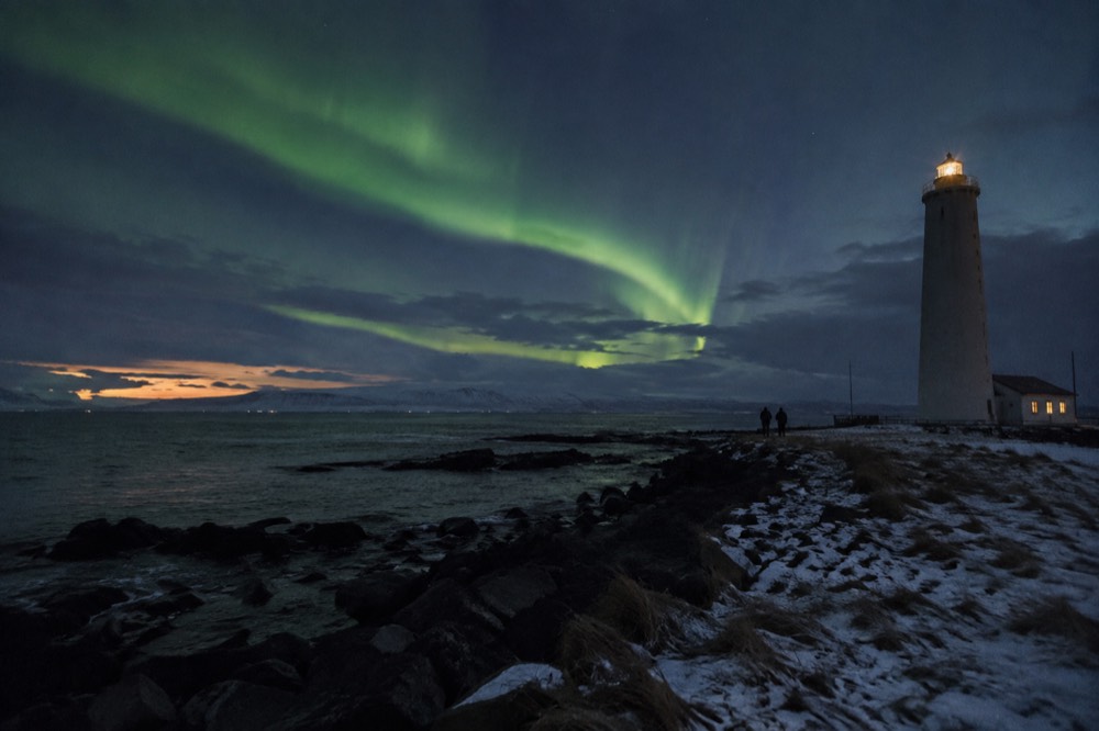 Green aurora arcs over a dark sea horizon with a lighthouse silhouette at the edge of the frame