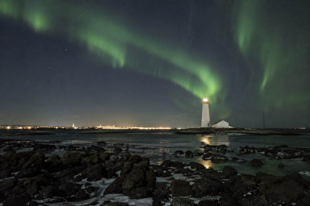 Aurora glow over a dark shoreline with a lighthouse silhouette