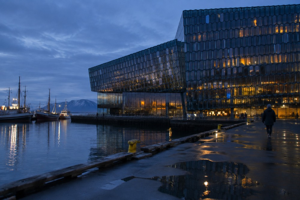 A concert hall facade of geometric coloured glass panels beside a harbour at dusk
