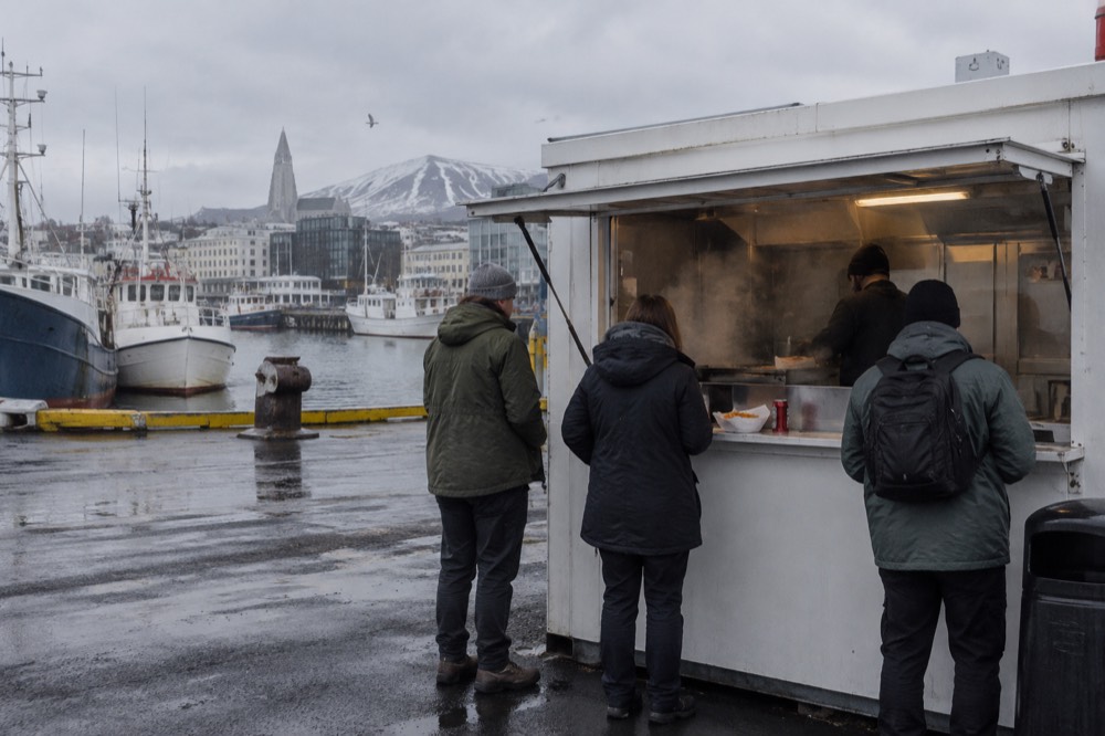 A small hot dog stand beside a harbour with a queue of customers