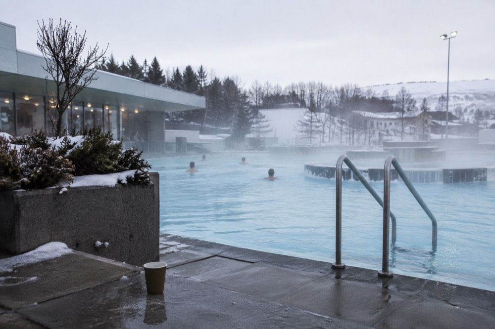 Outdoor geothermal pool with steam drifting across the surface and potted plants at the edge