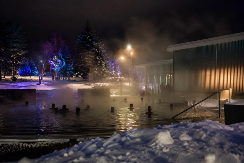 Illuminated outdoor pool at night with steam rising and festival lights on surrounding buildings