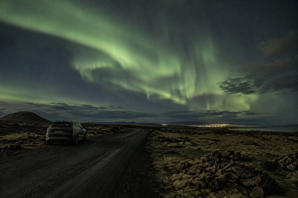 Wide view of aurora curtains over a dark hillside road outside the city