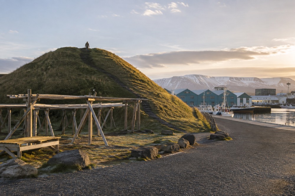 The grassy cone of the Thufa mound rising beside harbour buildings under a wide sky
