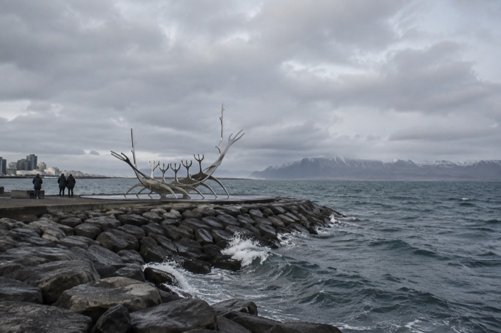 A curved steel boat skeleton sculpture facing the open water of a bay, with low hills on the far shore
