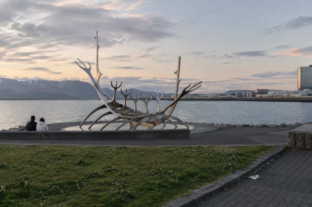 The steel boat sculpture seen from the side, with a paved waterfront walkway, grass and the bay beyond