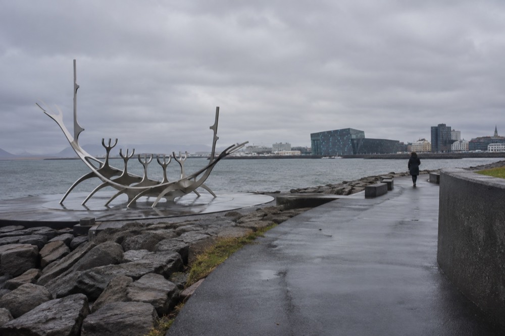 A paved waterfront path curving along a bay, with a concert hall of glass panels visible in the distance