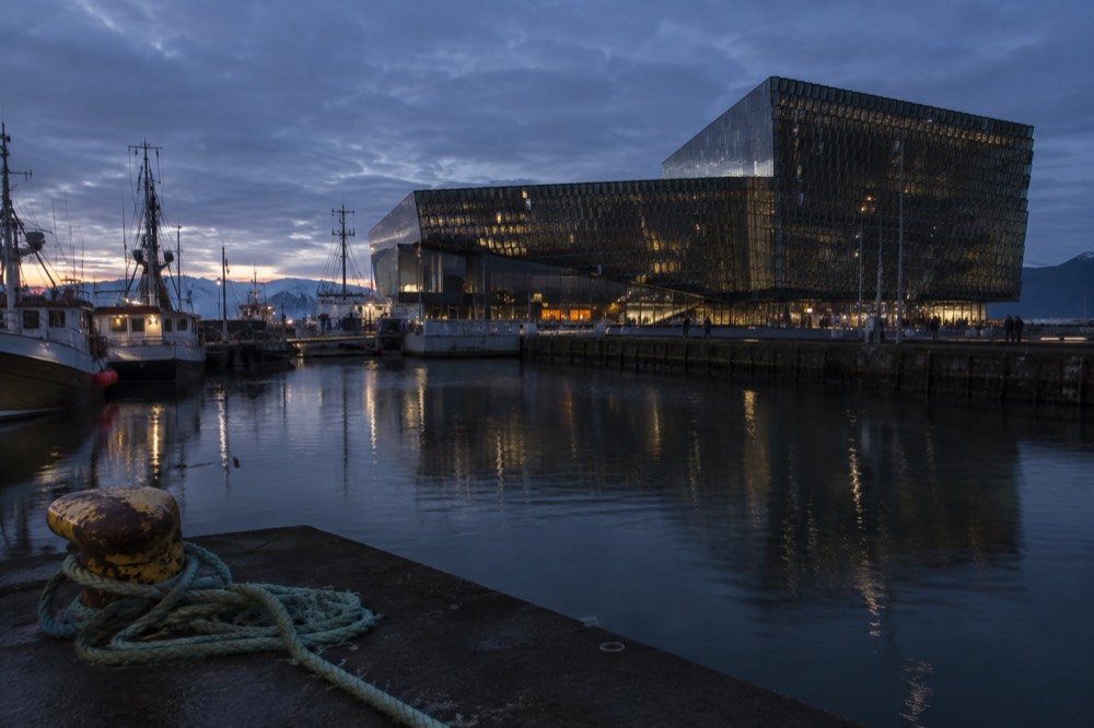 A glass-fronted concert hall with faceted windows beside a harbor, with small boats moored nearby