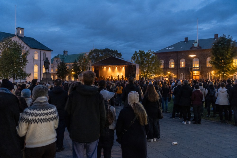 A crowd gathered in a city square at dusk with stage lights